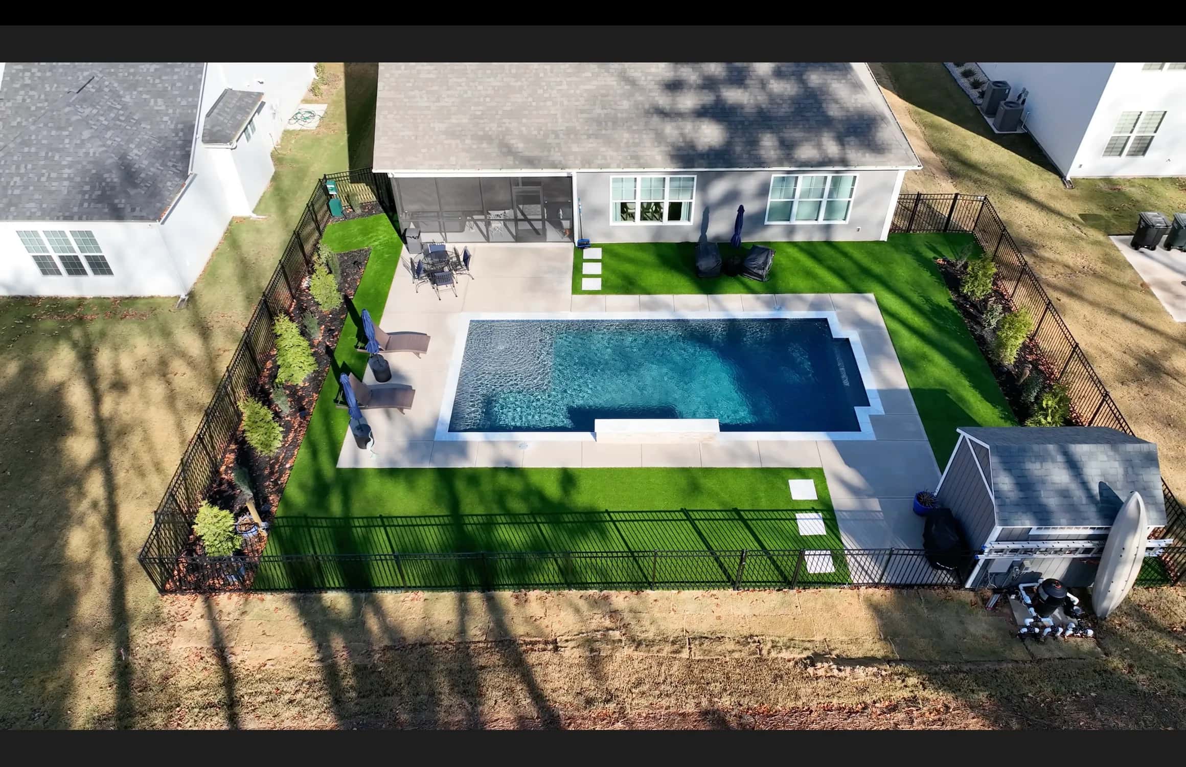 An elevated photograph shows a rectangular pool surrounded by a broad light-colored deck and planted edges.