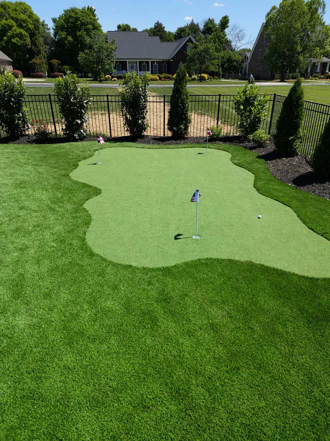 A backyard putting green with multiple flagsticks sits inside a larger green area beside perimeter planting.