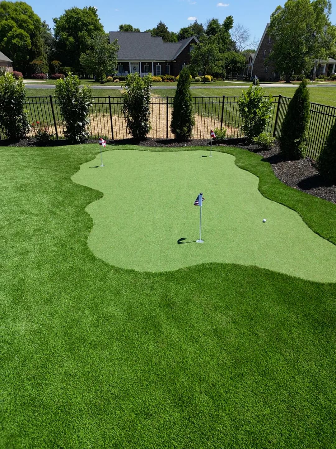 Three-hole putting green with a darker turf fringe and planting along a black fence
