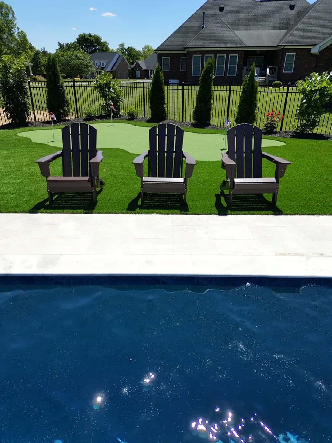 Three outdoor chairs sit on a green surface behind a pool deck, with a putting area and planting along the fence.
